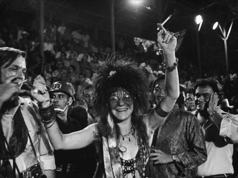 A imagem está em preto e branco. Janis Joplin, uma mulher branca, está no centro, com os braços levantados e um cigarro em uma mão. Ela usa regata, plumas na cabeça e pulseiras e colares. Ao seu redor, muitas pessoas curtem o carnaval do Rio de Janeiro.
