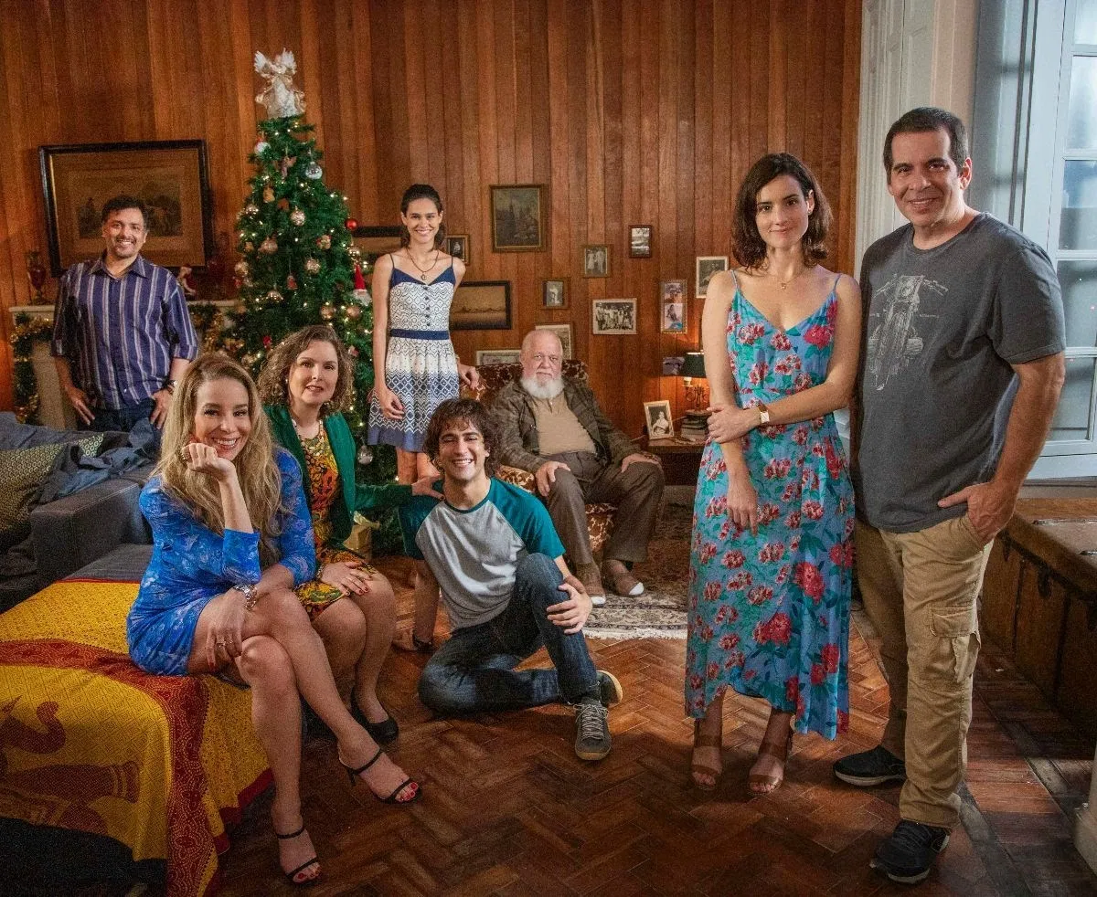 Imagem do filme Tudo Bem no Natal que Vem. Nela vemos 8 personagens, todos brancos, reunidos na sala de uma casa, com paredes de madeira com alguns quadros pendurados e uma árvore de Natal no canto esquerdo.