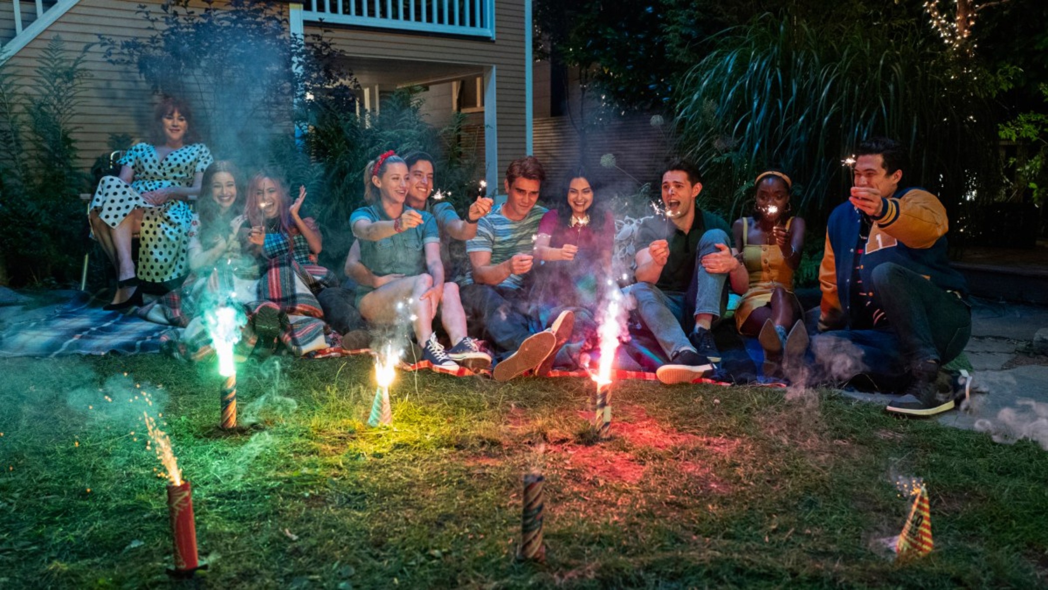 Cena da cena Riverdale. Na cena, vemos uma porção de pessoas sentadas num gramado, sorrindo, enquanto fogos de artifício queimam à frente deles. 
