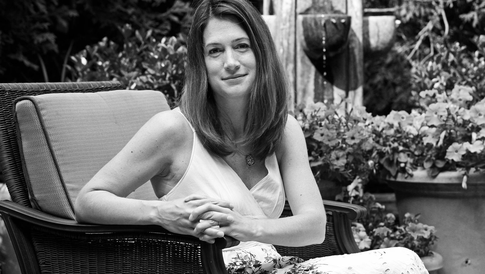 Foto em preto e branco da autora Gillian Flynn. Ela é uma mulher de 50 anos branca com cabelos castanhos na altura do ombro, usando um longo vestido branco sem mangas. Gillian Flynn está sentada em uma poltrona de vime, com as mãos entrelaçadas e algumas plantas no fundo. Olha diretamente para a câmera, e está sorrindo sem mostrar os dentes.