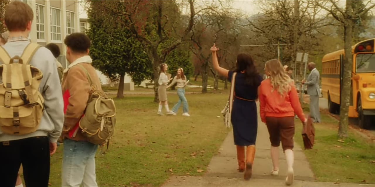 Cena da série Amigas Para Sempre, print de tela, Tully e Kate, duas mulheres brancas, ainda adolescentes caminhando em frente à escola, as duas estão de costas e Tully mostra o dedo do meio. A foto tem a temperatura mais quente