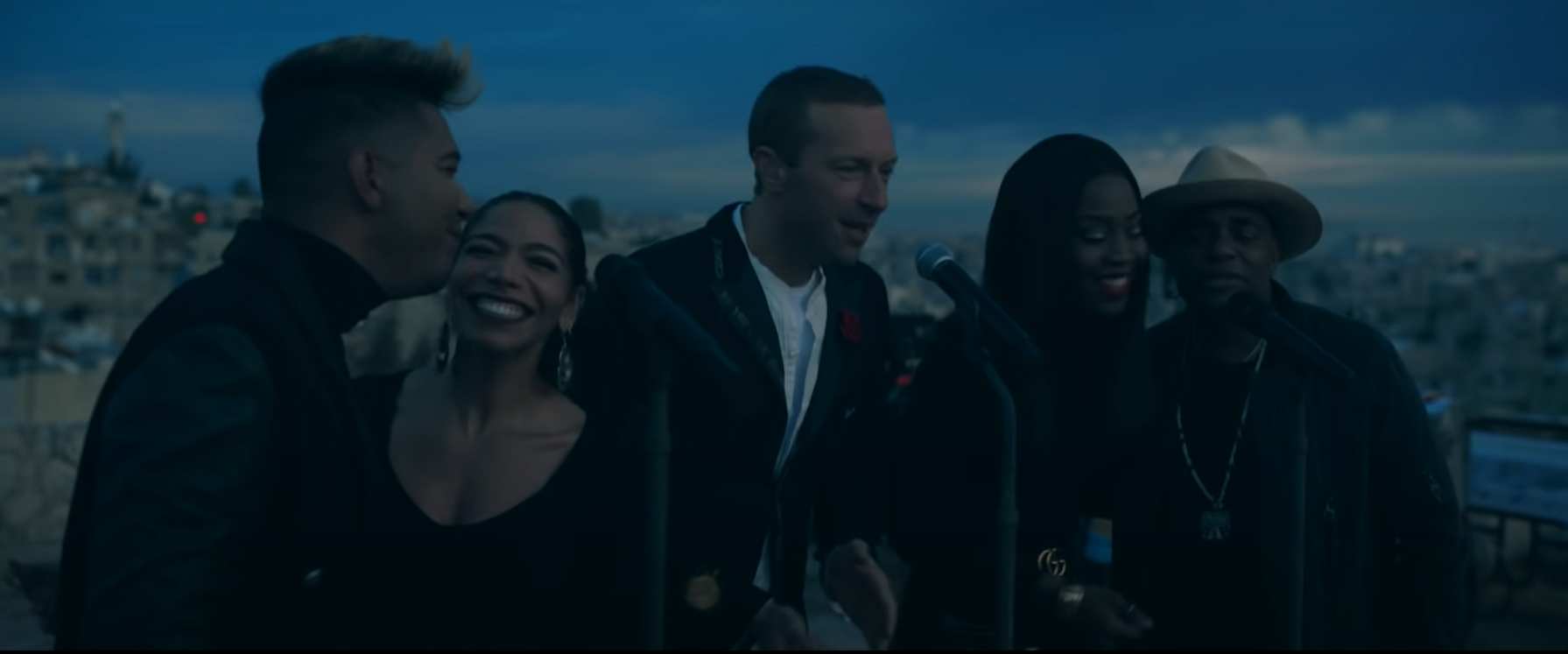 Imagem da apresentação que Coldplay fez ao vivo do disco Everyday Life. Na fotografia, o vocalista Chris Martin está ao centro, na frente de um microfone, cantando. Ao lado esquerdo dele, estão dois cantores, primeiro, à esquerda da imagem, um homem negro de cabelo liso e logo depois uma mulher, também negra, de cabelos presos, batom escuro e sorridente. Eles dividem um microfone, que está logo na frente deles. Então, à direita de Chris Martin, estão mais outros dois cantores. Primeiro, uma mulher negra, de cabelos lisos soltos, e depois, na extremidade direita da imagem, um homem, que usa um chapéu marrom. Eles também dividem um microfone e sorriem enquanto cantam. Ao fundo da imagem, está a cidade e o céu, ainda meio escuro por conta do amanhecer.