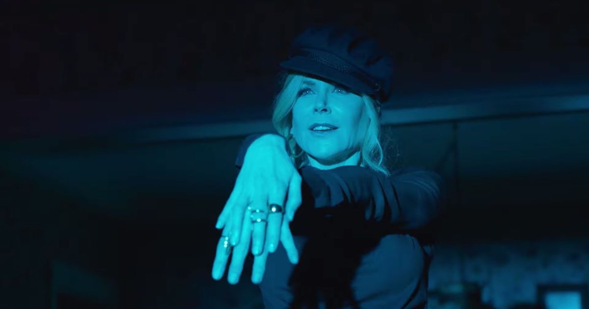 Cena do filme A Festa de Formatura, mostra Nicole Kidman com as mãos uma em cima da outra, estendidas a frente do corpo. Ela usa uma boina e blusa de mangas compridas, ambos pretos, tem os cabelos loiros presos com a franja solta na fente do rosto. Ela é uma mulher branca e está sendo iluminada por uma luz azul.