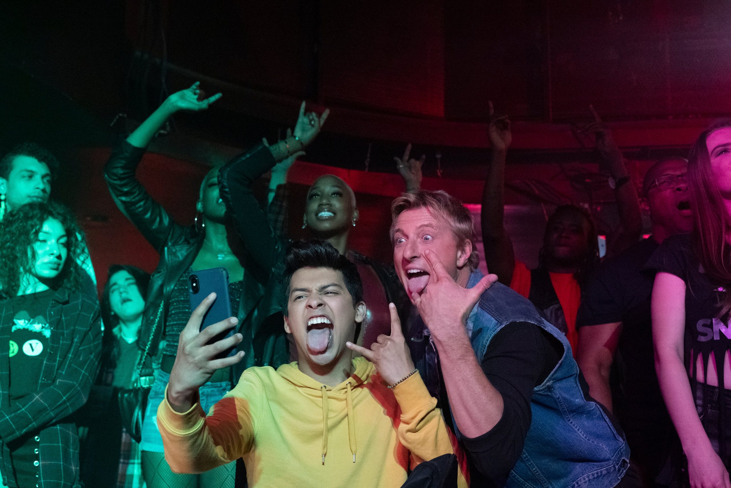 Dois amigos ao centro tirando uma selfie, fazendo sinal de rock n’ roll com as mãos e mostrando a língua. Ao fundo, um grupo de pessoas aproveitando um show de música 