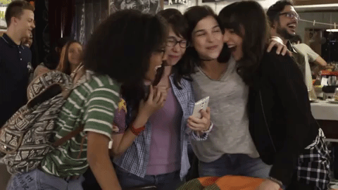 Na imagem em gif, as atrizes de As Five mais jovens, de quando estavam em Malhação: Viva a Diferença, se abraçam. Ao fundo, um restaurante com alguns figurantes em pé.