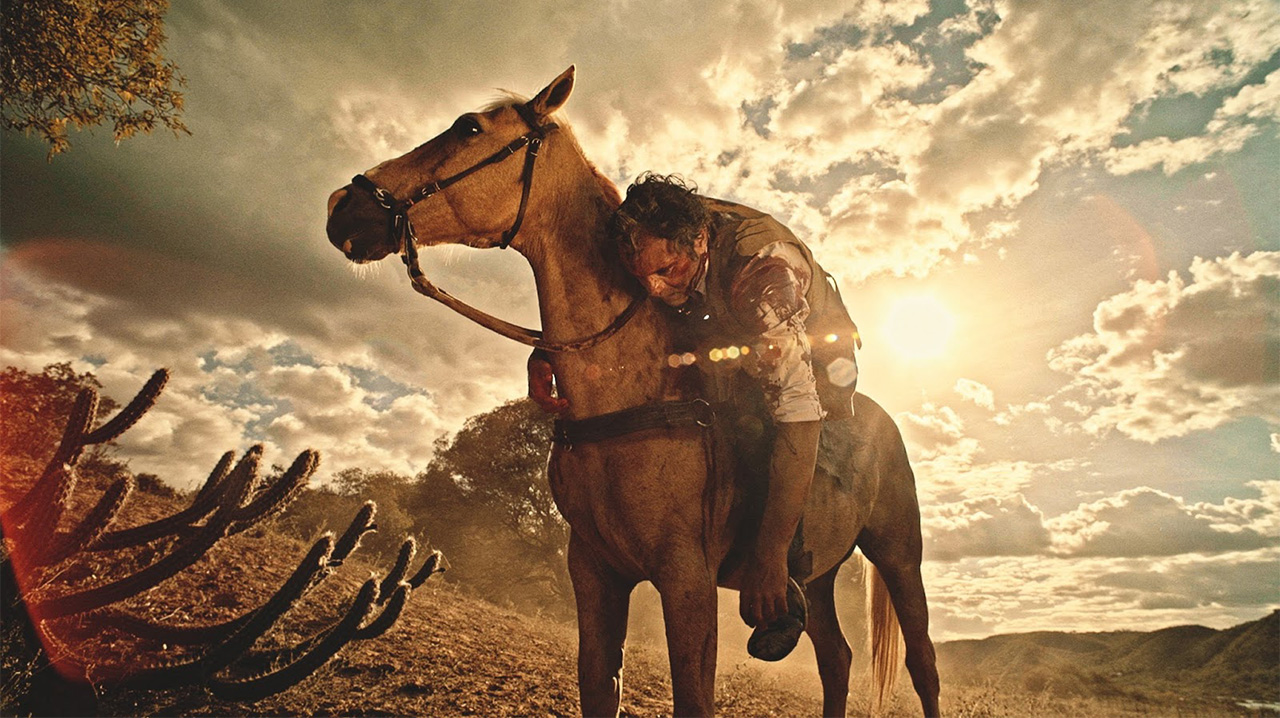Cena da novela Velho Chico. Santo, enquanto cavalga, é alvejado por um tiro no ombro. No fundo, vemos o céu luminoso, entre nuvens, e a paisagem da caatinga.