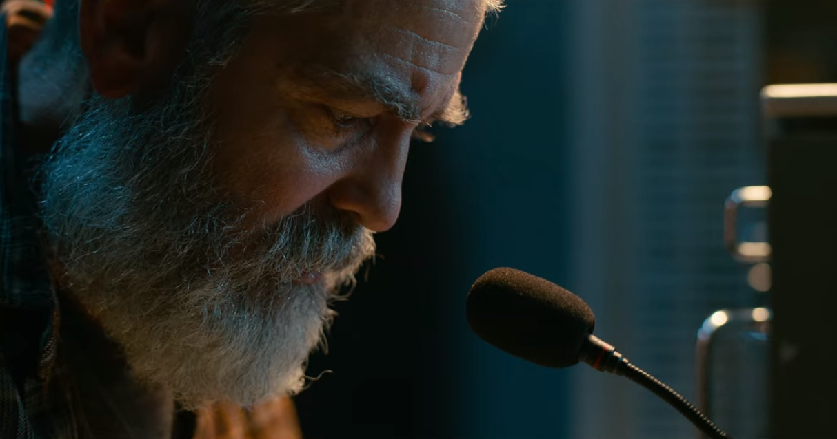 Cena de O Céu da Meia Noite. Vemos o rosto de George Clooney bem próximo, de perfil. Ele interpreta Augustine e está de frente para um microfone preto. Uma luz suave e amarelada em seu rosto contrasta com as sombras e o fundo azul escuro.