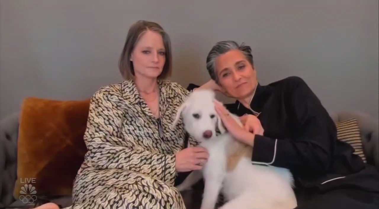 Foto da atriz Jodie Foster. Uma mulher branca com cabelos até a altura do ombro, e está sentada em um sofá cinza com almofadas laranjas do lado de um cachorro branco e outra mulher branca. Ambas vestem pijamas, jodie um conjunto claro e sua mulher um conjunto preto.