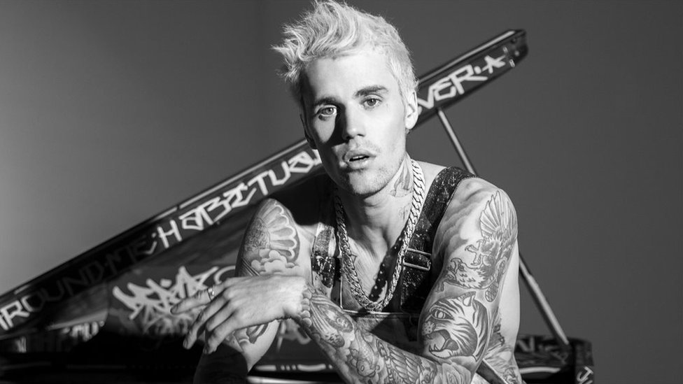 A imagem é uma fotografia do cantor Justin Bieber, em preto e branco. Nela, Justin está sentado com o braço esquerdo apoiado em sua perna, olhando fixamente para a câmera. Justin é um homem branco, de cabelos lisos e com várias tatuagens no corpo, ele está sem camisa e usando uma corrente no pescoço.