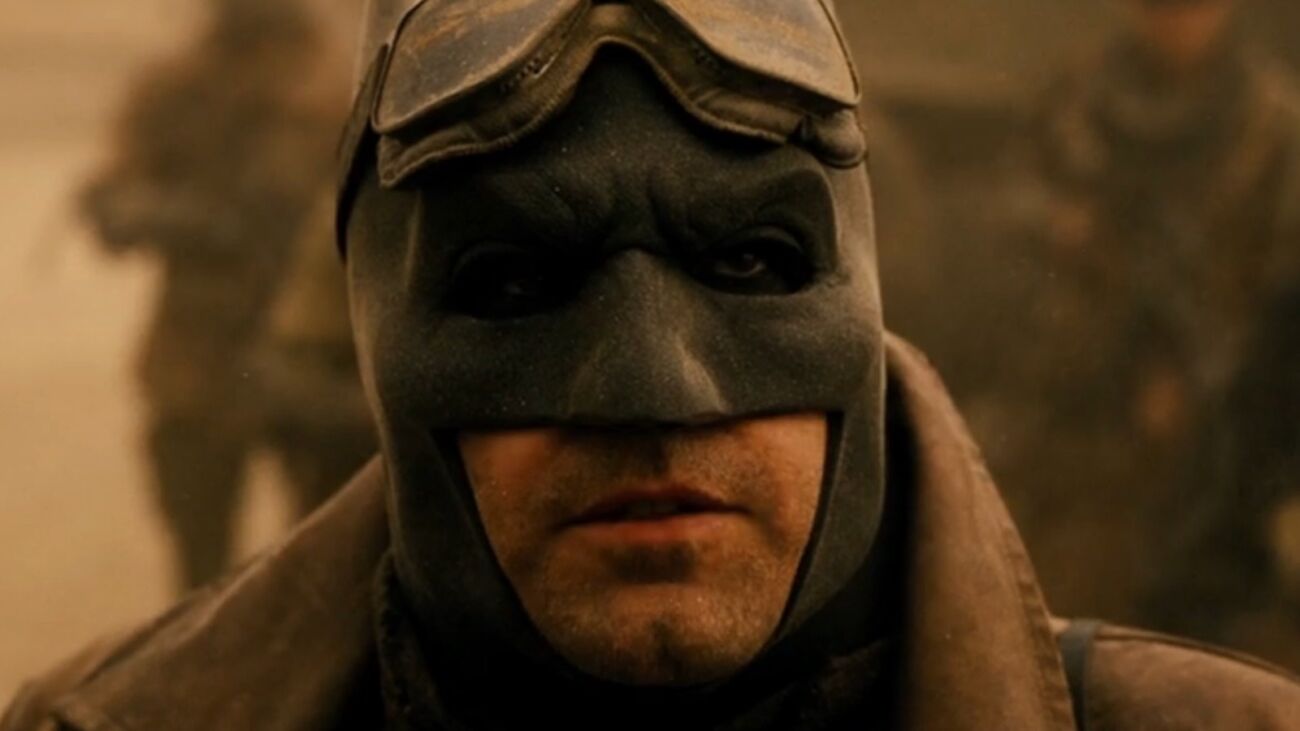 Cena do Snyder Cut. Na cena, vemos um close-up do Batman. Ele tem pele branca, e veste a máscara preto de borracha em formato de morcego. Acima da máscara, usa um óculos de aviador que está posicionado acima de seus olhos, na testa. Ele veste uma japona bege e o cenário é desértico.