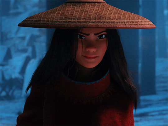 A personagem principal do filme, Raya, está usando um manto vermelho, com um chapéu de palha por cima de seu cabelo solto. Ela encara a câmera com uma expressão determinada e sorri de canto