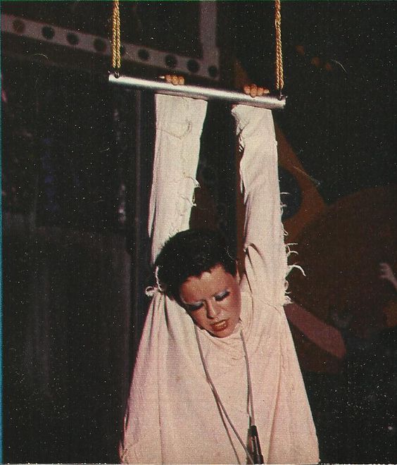 Fotografia de Elis Regina no espetáculo Falso Brilhante. Na imagem, a artista segura uma barra com as mãos e parece estar pendurada. Ela olha para baixo, com os olhos quase fechados, e veste uma roupa branca. 