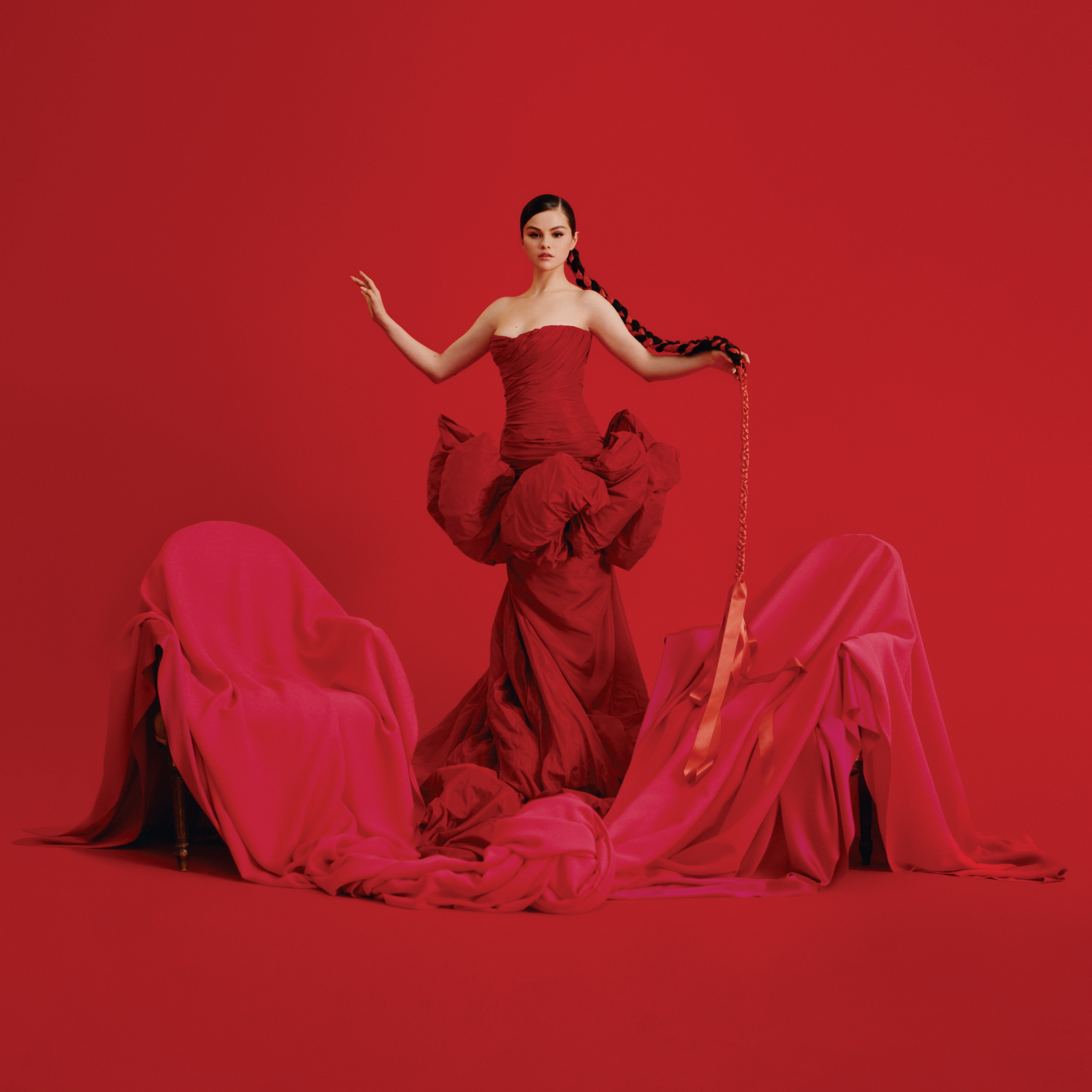 Selena Gomez, uma mulher branca de cabelo castanho, está em pé, usando um vestido vermelho com babados na cintura. Ela está com uma trança no cabelo. Essa trança está com uma fita vermelha, que vai até as pontas do cabelo e com um caimento de fita abaixo. No chão, existem duas cadeiras, cobertas por um pano vermelho. O fundo da imagem também é todo vermelho.