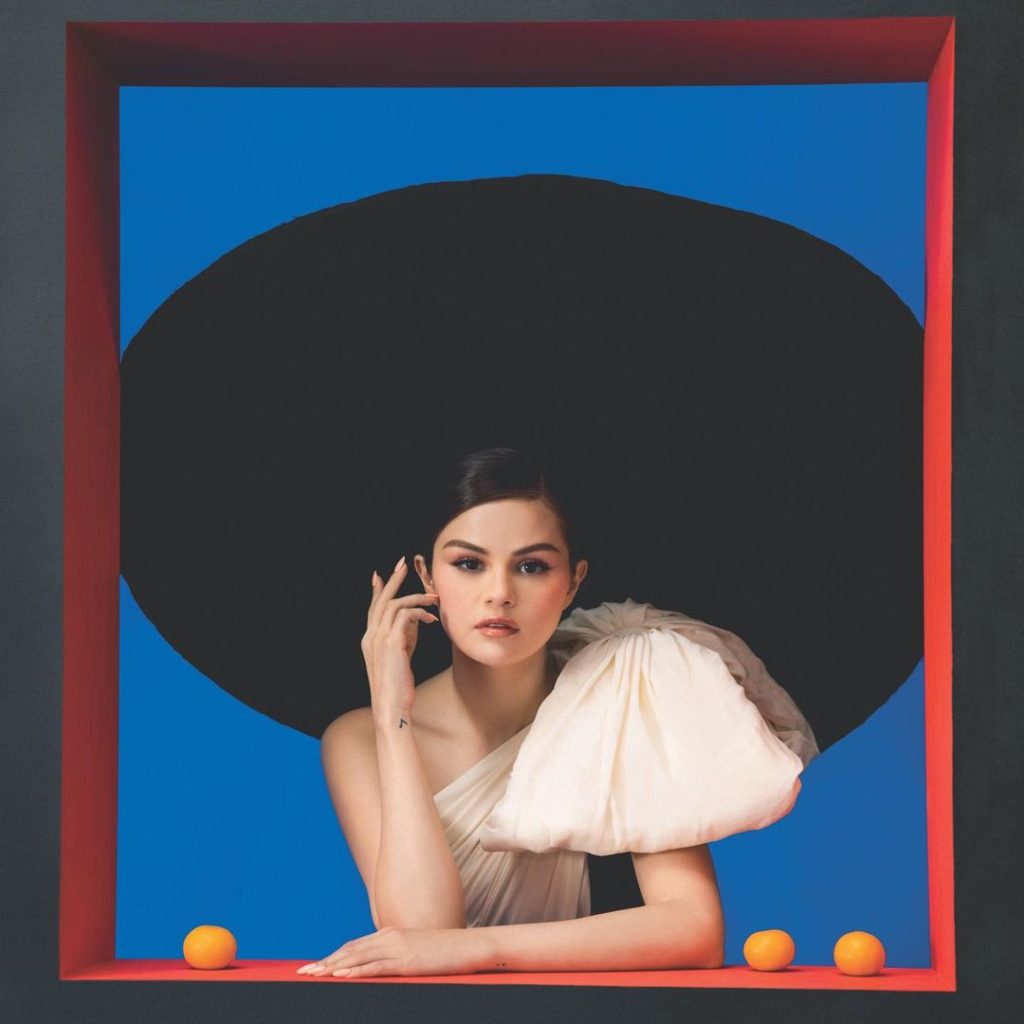 Selena Gomez está com o cabelo preso, usando um chapéu preto gigante, com um vestido bege de um ombro só. O vestido tem um laço gigante no ombro. Ela está com a mão no queixo e ao seu lado têm três laranjas, duas à direita e uma à esquerda. Em seu braço existe uma tatuagem de nota musical. O fundo da imagem é azul, com uma borda vermelha, parecendo uma janela ou um quadro.
