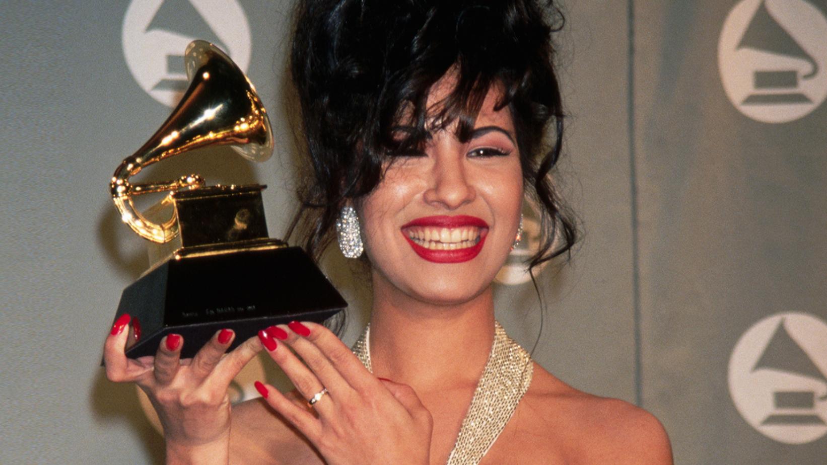 Selena Quintanilla, uma mulher branca de cabelo preto, está segurando seu gramofone dourado. Ela está de cabelo preso, com a franja no rosto, brincos brilhantes e unhas grandes pintadas de vermelho. Ela está sorrindo, com um batom vermelho e a alça de seu vestido é brilhante. Na mão em que está segurando o Grammy, ela usa um anel prata.