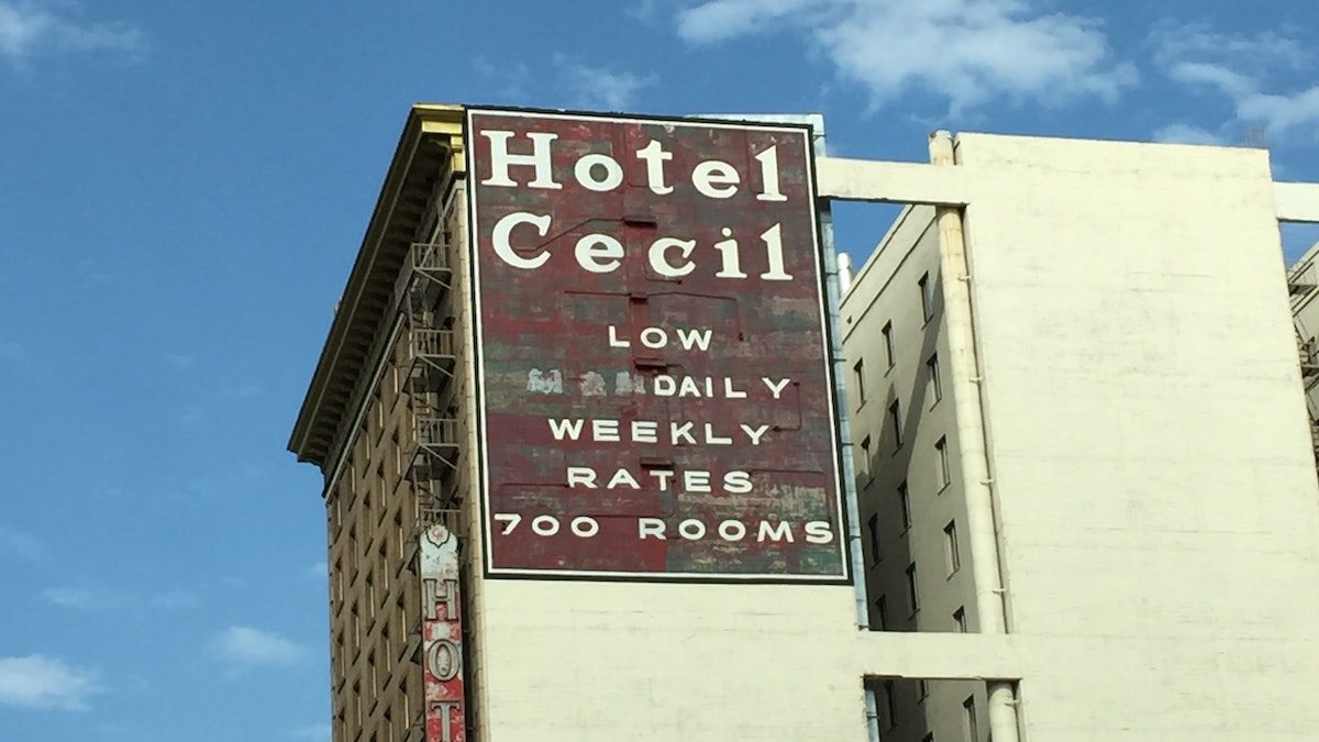 Foto do Hotel Cecil. O hotel é bege, com uma grande placa vermelha no que anuncia HOTEL CECIL - LOW DAILY WEEKLY RATES - 700 ROOMS. A fachada está desgastada e o céu no fundo é azul.