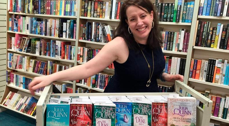 A autora Julia Quinn, mulher branca de olhos e cabelos castanhos, sorrindo mostrando seus livros da série Os Bridgertons na estante numa livraria. Ela usa uma blusa preta sem mangas, um colar dourado comprido e brincos de argola. Ao fundo vemos vários livros.