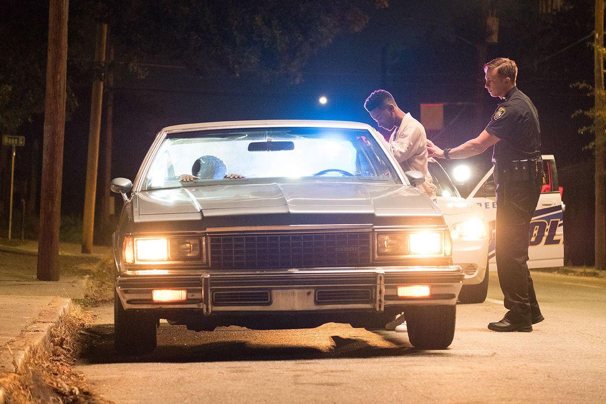 Foto de Khalil sendo abordado pelo Policial. Ele esta virado para seu carro, um impala cinza com os faróis ligados, enquanto o Guarda branco com seu uniforme o revista.