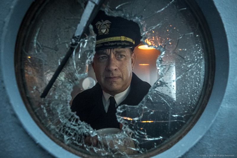  Cena do filme Greyhound. Nela vemos Tom Hanks, um homem branco e de meia-idade, com roupa de capitão. Ele olha por uma janela de navio quebrada.