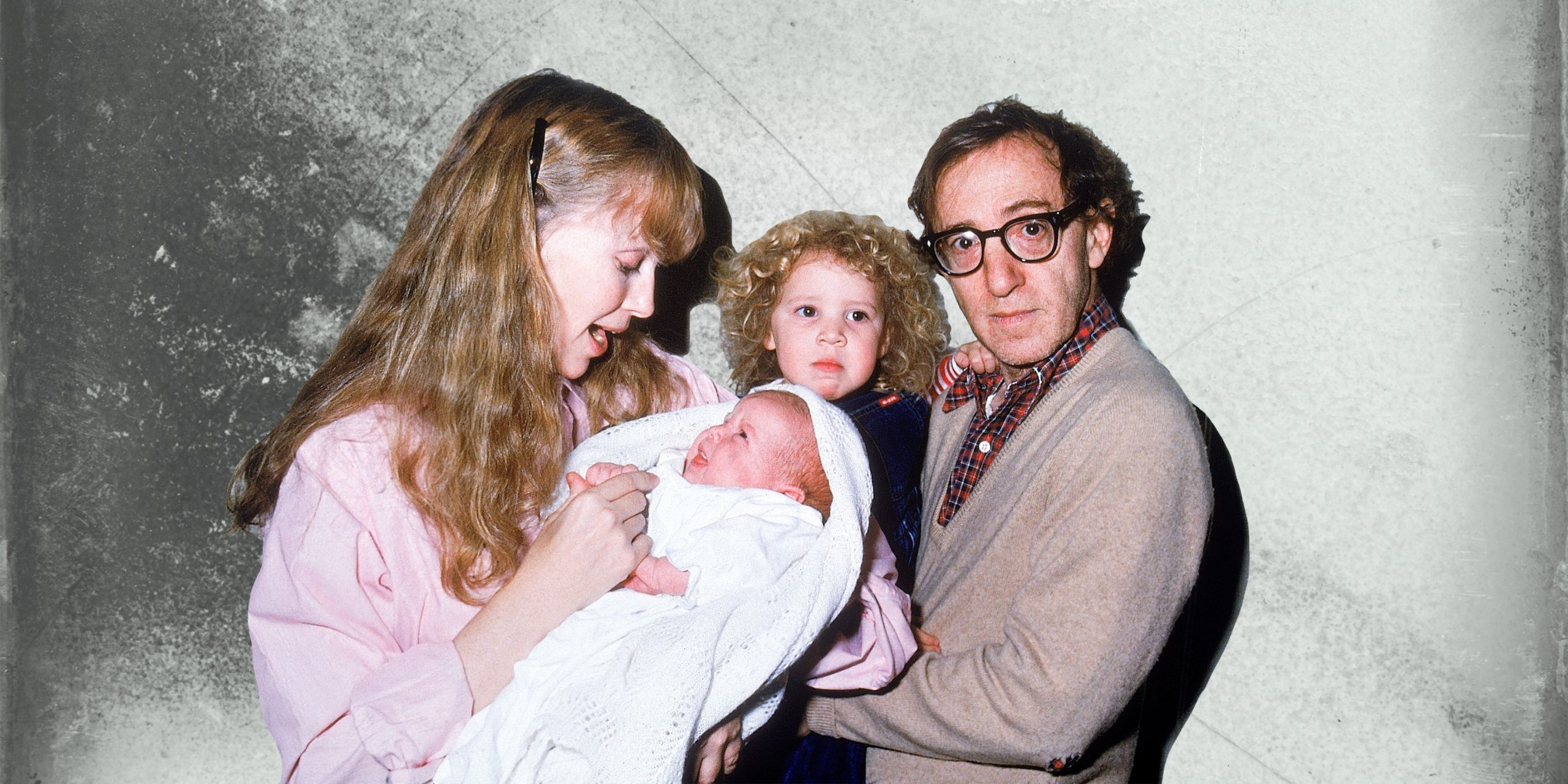Cena da série documental Allen v. Farrow. Nela, vemos uma fotografia antiga de Woody Allen, branco, idoso, de cabelos pretos e óculos redondos com armação escura, com sua filha Dylan no colo. Ao seu lado, está sua esposa Mia Farrow, branca, loira, segurando o filho recém-nascido no colo. 