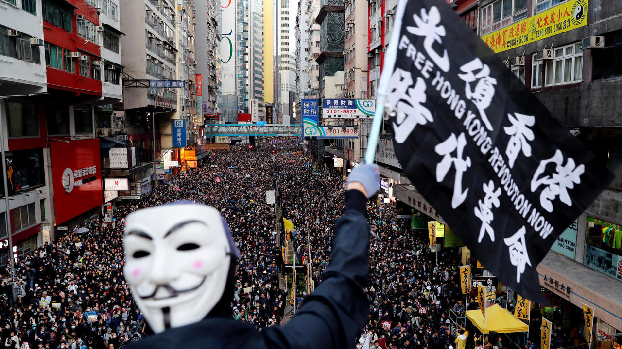 Fotografia de um protesto na cidade de Hong Kong. Em primeiro plano vemos um manifestante usando uma máscara do Guy Fawkes. Ele segura uma bandeira com os escritos FREE HONG KONG - REVOLUTION NOW. Ao fundo, podemos ver uma rua com milhares de manifestantes, preenchida de calçada a calçada. Aos lados, diversos prédios altos e fachadas de lojas. 