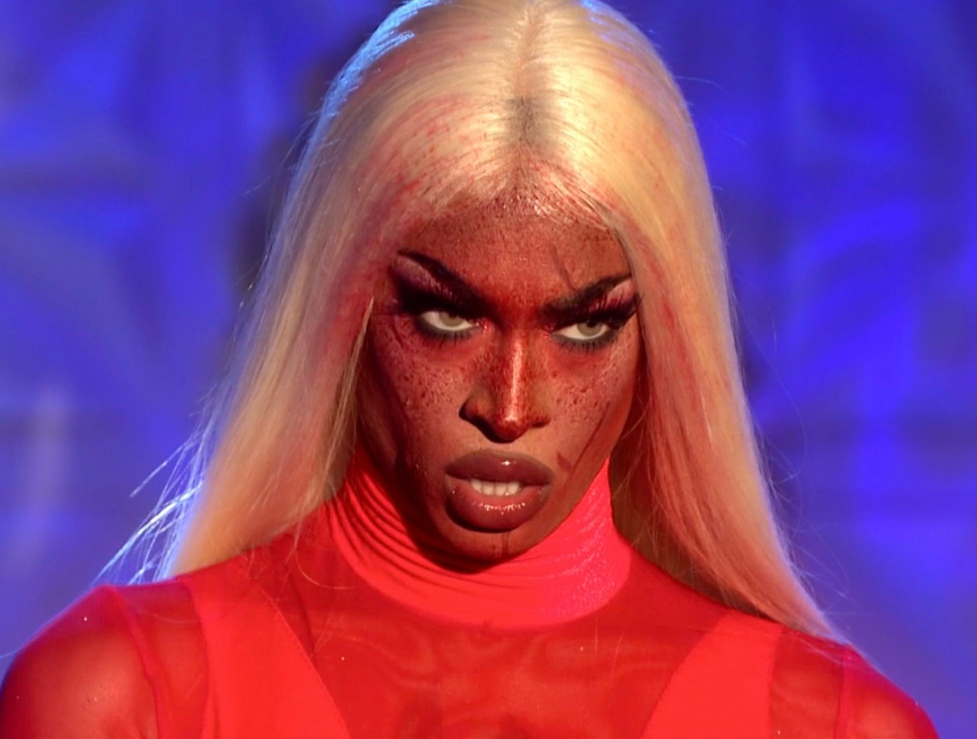 Cena de uma Dublagem da 2ª temporada de Drag Race UK. Nela, vemos um close no rosto de Tayce, uma drag queen negra, loira e com sangue no rosto. Ela veste vestido vermelho e olha para a frente, com um olhar determinado. 