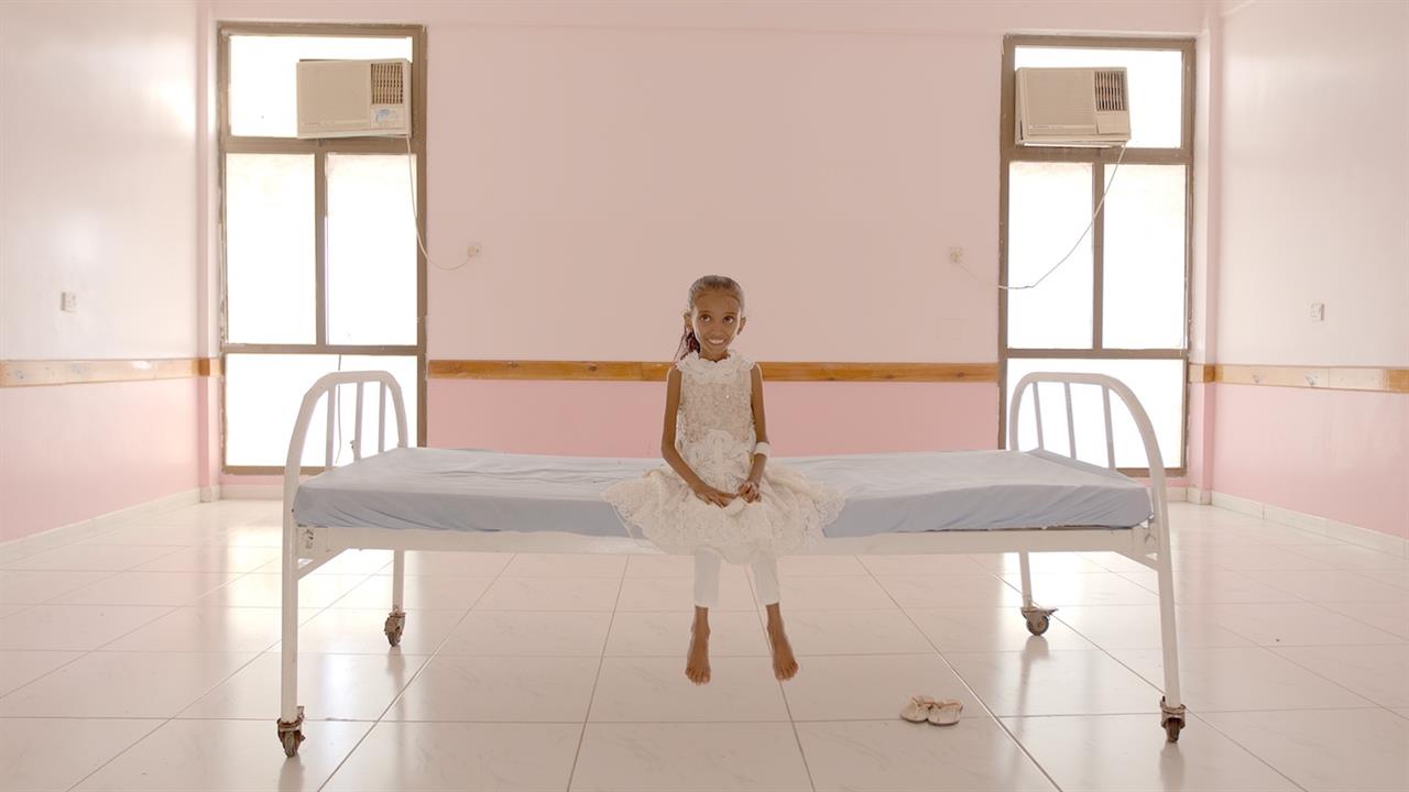 Foto de divulgação de Hunger Ward. Na imagem, uma menina iemenita, muito magra, aparentando ter cerca de cinco anos, de pele amarronzada e cabelos castanhos, senta sob uma cama, ao centro, e sorri para a câmera. Ela está em uma sala de paredes rosadas na clínica de reabilitação.