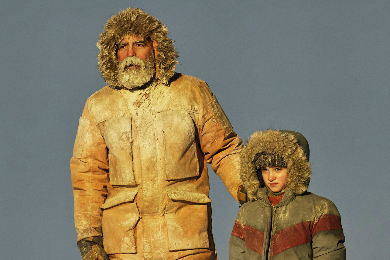 Cena do filme O Céu da Meia-Noite. Nela vemos George Clooney e uma criança. Eles estão vestindo casacos para neve e estão com neve em suas vestimentas. O céu ao fundo está limpo e o sol bate diretamente nas duas personagens, que olham para frente ao horizonte. George Clooney está com a barba congelada. 