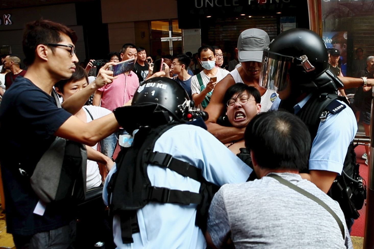 Cena do curta Do Not Split. Ao centro, vemos um grupo de policiais prendendo um manifestante honconguês. O manifestante está sendo segurado por um homem de boné com um mata leão. Pessoas em volta estão gravando a cena utilizando celulares. 