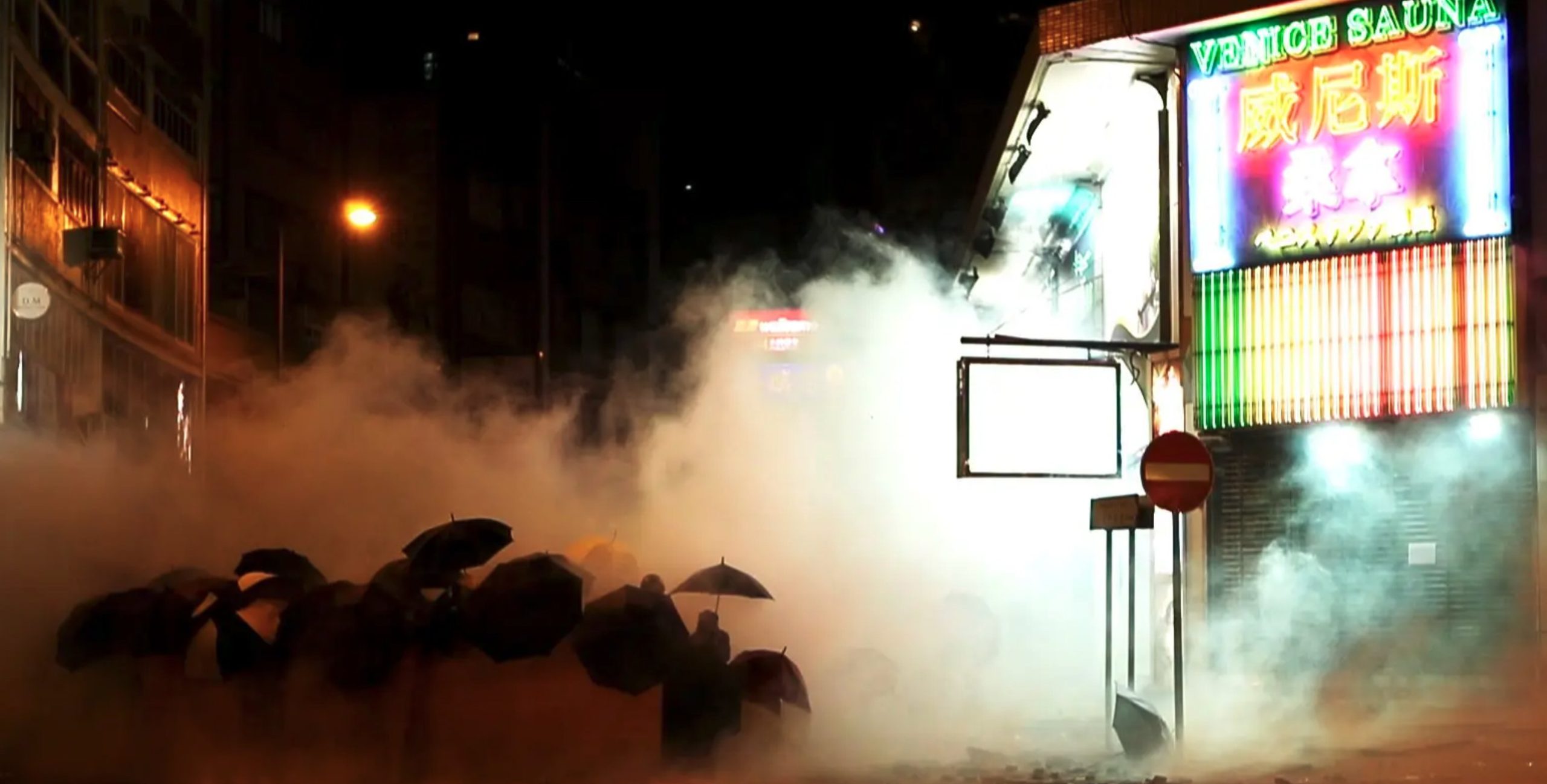 Cena do curta Do Not Split. Em meio a fumaça de gás lacrimogêneo, podemos ver vários manifestantes pró-Hong Kong segurando guarda chuvas. Eles estão em uma rua, é noite, e há um edifício comercial ao lado direito.
