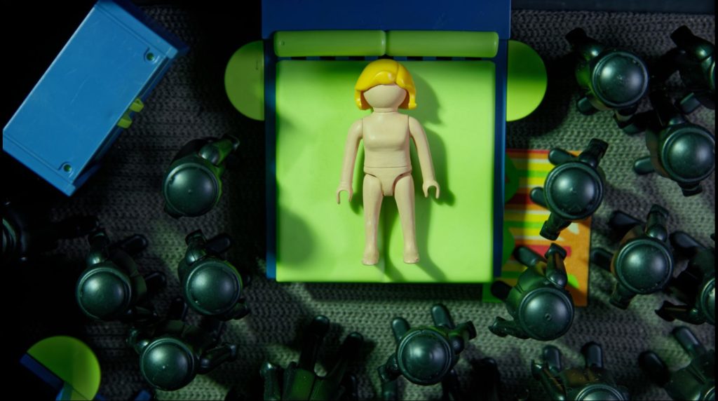 Cena da animação The Wedding Cake. Os personagens são todos bonequinhos de plástico. No centro, há uma mulher deitada em uma cama verde e azul. Ela é loira e está nua. Ao redor dela, vários bonecos pretos estão de pé na direção da cama. 