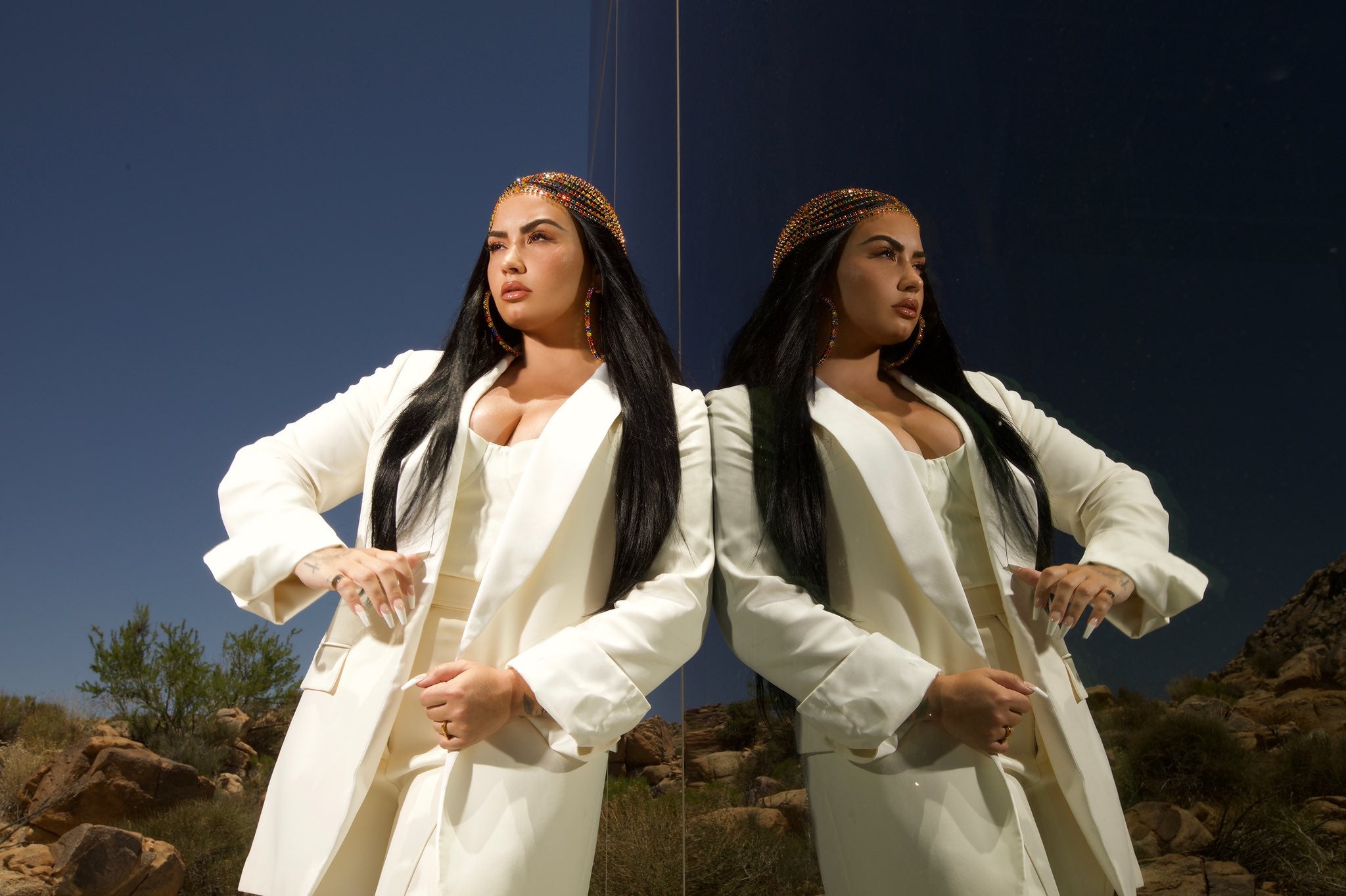 Foto de Demi Lovato. A cantora se encontra na frente de um espelho no deserto, onde sua imagem é refletida por completo no lado direito. Ela usa um blazer branco alongado até os joelhos e um macacão branco.