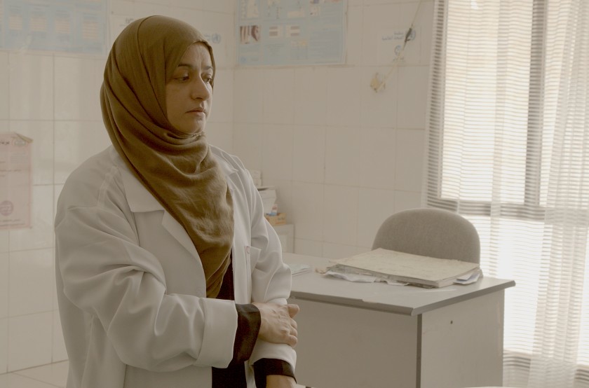 Cena de Hunger Ward. Na imagem, a médica Aida Alsadeeq, uma mulher de pele amarronzada, vestindo um jaleco branco e um hijab marrom, está em pé em frente a uma mesa branca, em seu consultório na clínica de reabilitação.