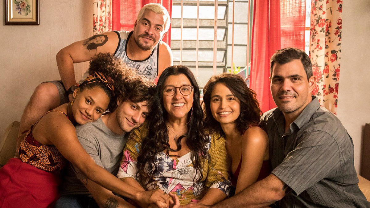 Cena da novela Amor de Mãe. Nela, vemos a família de dona Lurdes reunida no sofá da casa, sorrindo para a foto. À esquerda e atrás de todos, está Ryan, de cabelo descolorido e regata cinza. Abaixo dele, da esquerda à direita, vemos Camila, mulher negra vestindo roupa vermelha; Danilo, homem branco, de cabelos castanhos e camiseta cinza. Dona Lurdes está ao centro, sorrindo, de óculos com acessório que impede de cair, e cabelos pretos. Ao lado dela, estão Érica e Magno. 