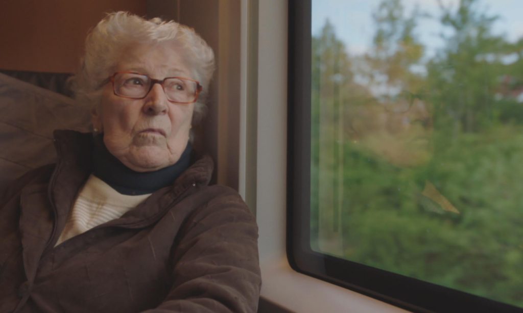 Cena do curta Colette.Nela vemos Collete, uma mulher de 90 anos, branca e com cabelo branco que veste um casaco marrom e está apoiada na janela de um trem. A vista é de várias árvores.