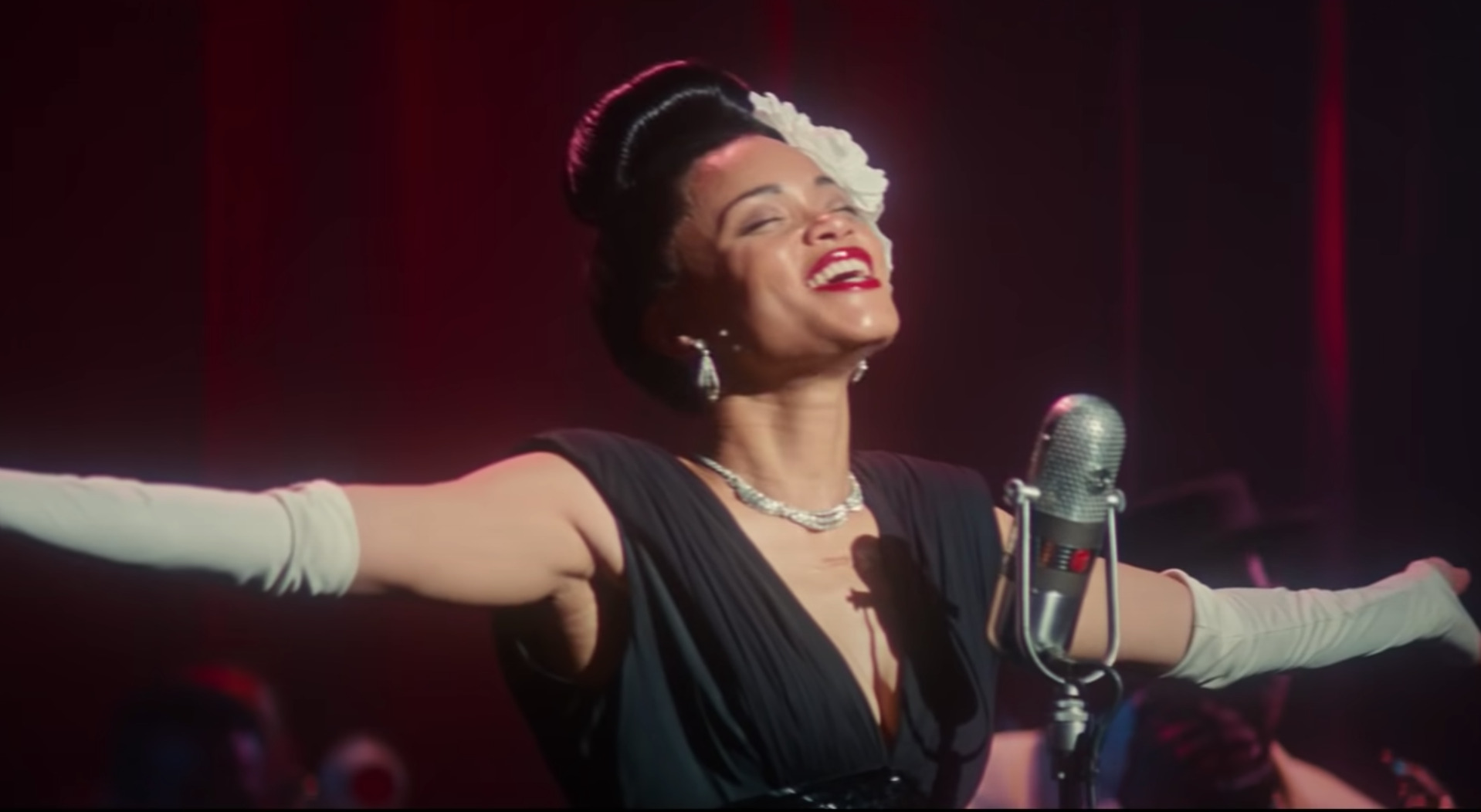 Cena do filme Estados Unidos Vs Billie Holiday. Na imagem, vemos Billie Holiday, interpretada por Andra Day, em frente a um microfone, com os braços abertos. Ela é uma mulher negra, de cabelos escuros presos em um coque no topo da cabeça, com uma flor branca ao lado esquerdo. Billie veste um vestido preto, luvas brancas compridas, um par de brincos e um colar. Ela está sorrindo e com os olhos fechados.