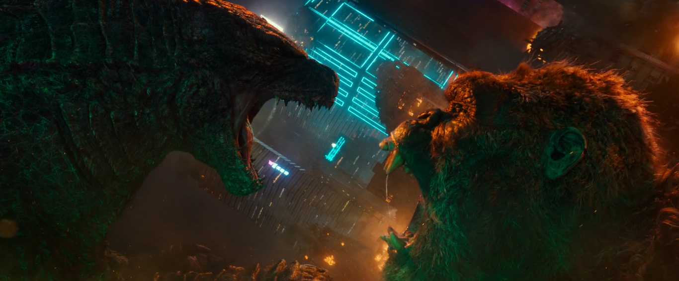 Cena do filme Godzilla vs. Kong. A imagem mostra os dois titãs, de perfil e na diagonal, encarando um ao outro. Eles estão de boca aberta mostrando os dentes. Godzilla está no canto superior esquerdo, e Kong está no canto inferior direito. Ao fundo, vemos edifícios da cidade de Hong Kong, à noite.
