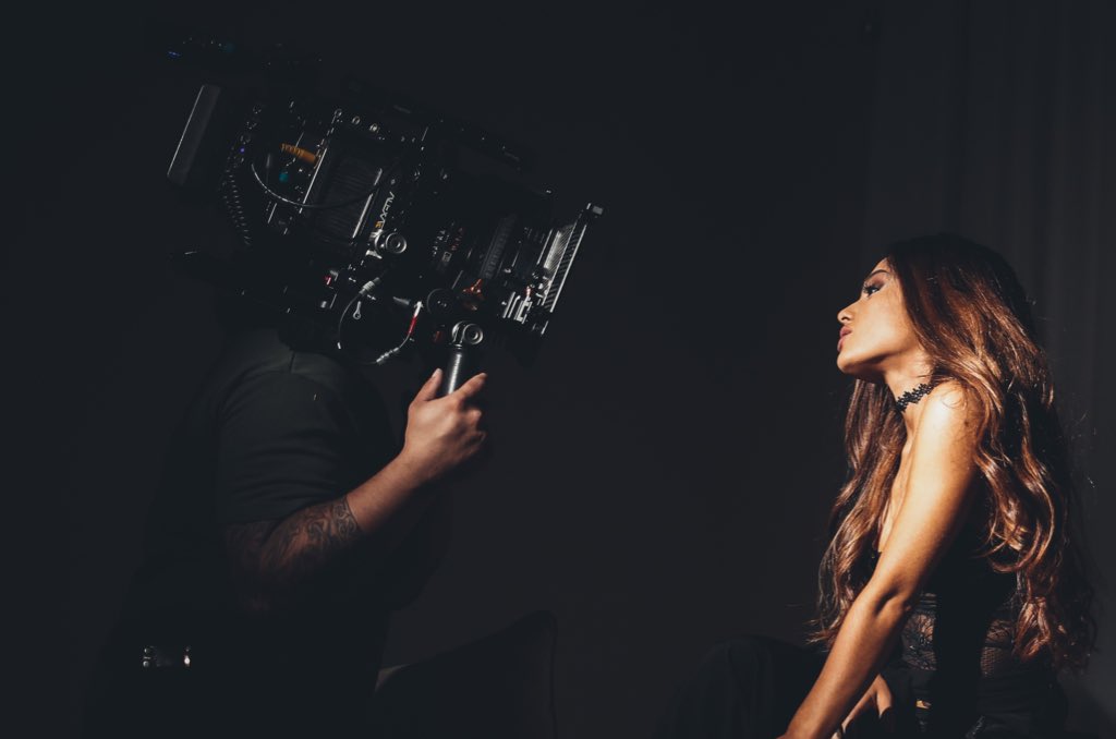 Gravação do clipe Dangerous Woman. Um diretor segura uma grande câmera na parte esquerda. Apenas seus braços aparecem. Ariana está virada, de perfil. Seus cabelos estão soltos e são longos. Ela veste uma lingerie preta.