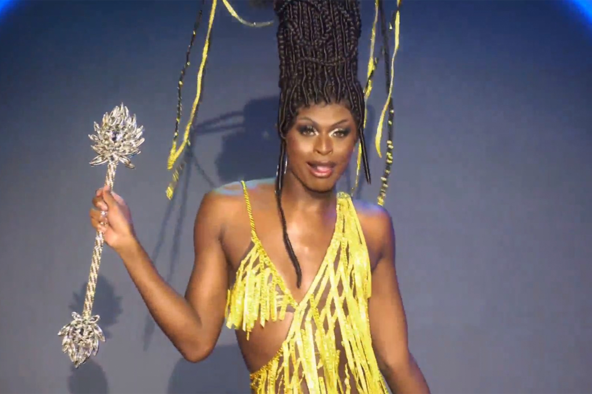 Cena da 13ª temporada do reality show RuPaul’s Drag Race. Nela, vemos Symone, drag negra, magra e de peruca preta com adornos de confete dourados, andando com um cetro prateado. Seu vestido segue o mesmo padrão amarelo dos confetes do cabelo