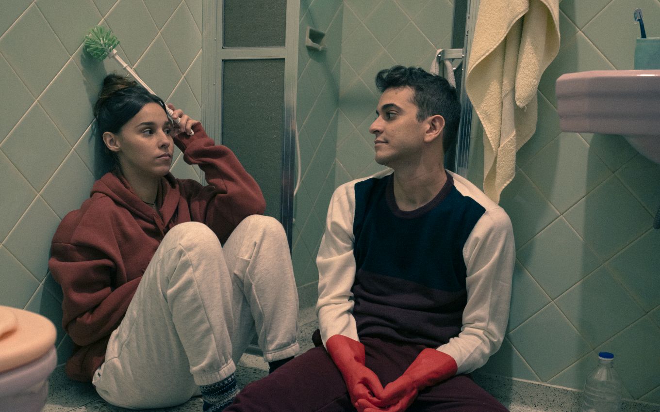 Cena do filme Me sinto bem com você. Nela, vemos o casal Thati e Victor. Eles são adultos, brancos, de cabelos escuros e estão sentados no chão do banheiro. Eles se olham e ela segura a escova de lavar a privada. 