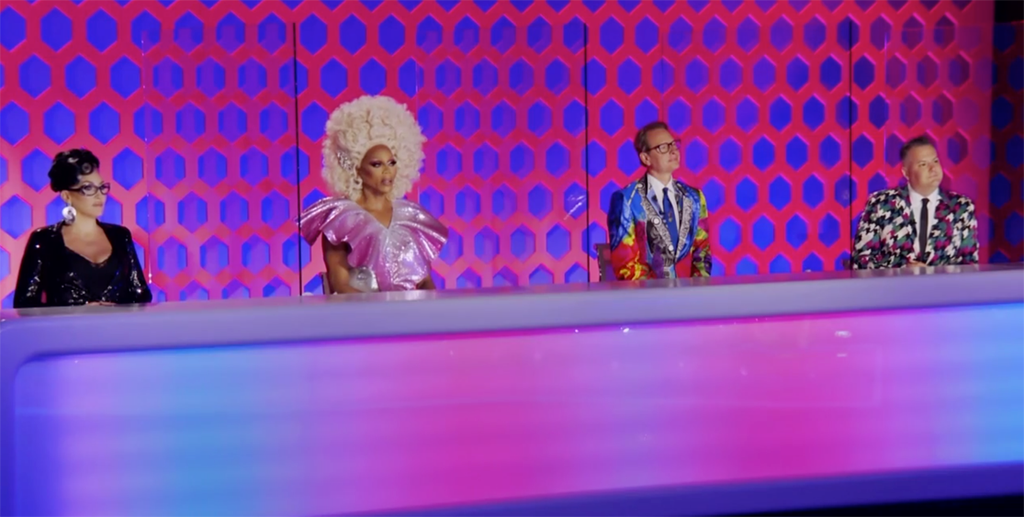 Cena da 13ª temporada do reality show RuPaul’s Drag Race. Nela, vemos o painel dos jurados, socialmente distantes. Da esquerda para a direita estão Michelle Visage, RuPaul, Carson Kressley e Ross Mathews.