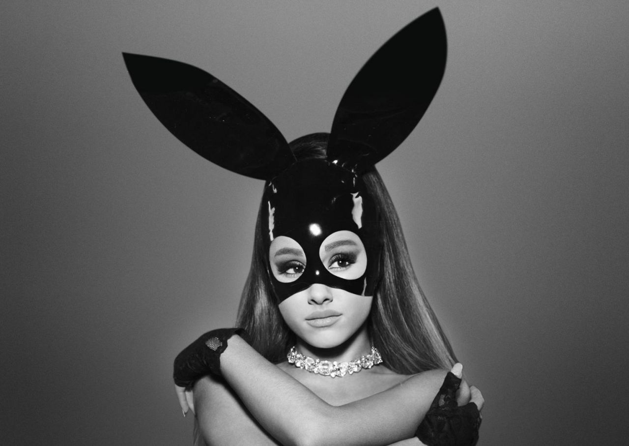 Ariana Grande se revela na imagem, de tronco para cima, abraçando seus ombros. A imagem está em preto e branco. Ariana é uma mulher branca, de cabelo castanho claro. Ela usa um vestido de látex preto e uma máscara preta com orelhas de coelho. Sua expressão é séria.