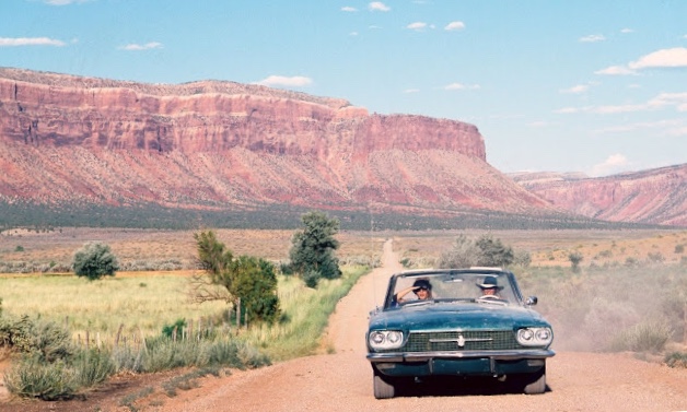 Cena do filme em que Thelma e Louise cruzam o deserto dentro de um carro conversível azul. A estrada é de terra, nos cantos está a vegetação baixa típica de clima desértico e uma montanha toma conta do fundo da imagem.