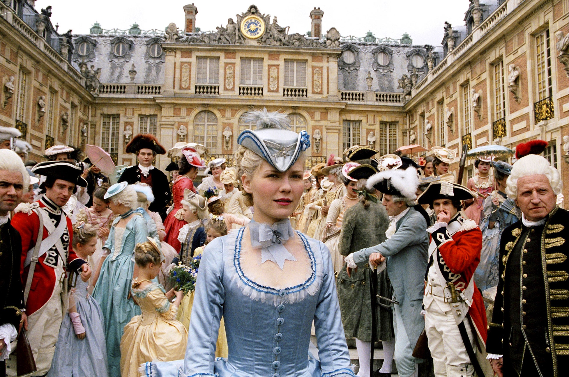 Cena do momento em que Maria Antonieta chega a Versalhes. Ela usa um traje azul claro, que contrasta com as roupas majoritariamente escuras da multidão que a cerca. Todos estão fora do palácio, cercados pela construção como se estivessem em um pátio.