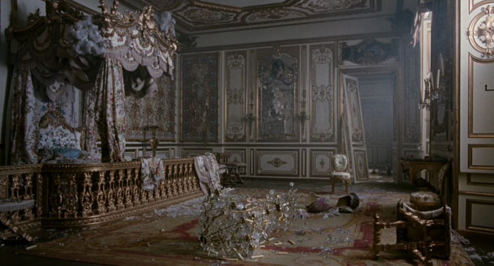 Cena do filme Maria Antonieta. Mostra um dos quartos do palácio totalmente destruído pelos revoltosos. Há um lustre de cristal despedaçado no chão, portas quebradas e mobílias reviradas. A imagem possui um tom acinzentado.