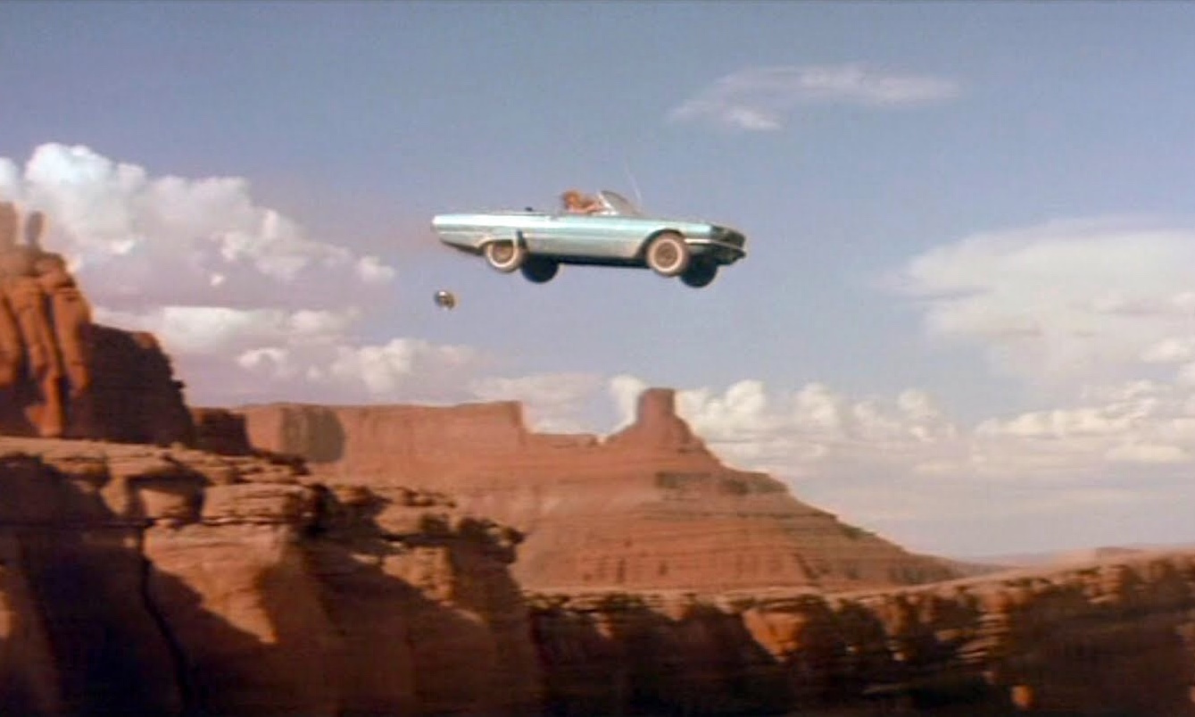 Cena do filme em que um carro azul conversível sobrevoa o Grand Canyon. A metade para baixo da imagem é composta por rochedos desérticos e a parte superior é o céu azul com poucas nuvens dispersas.