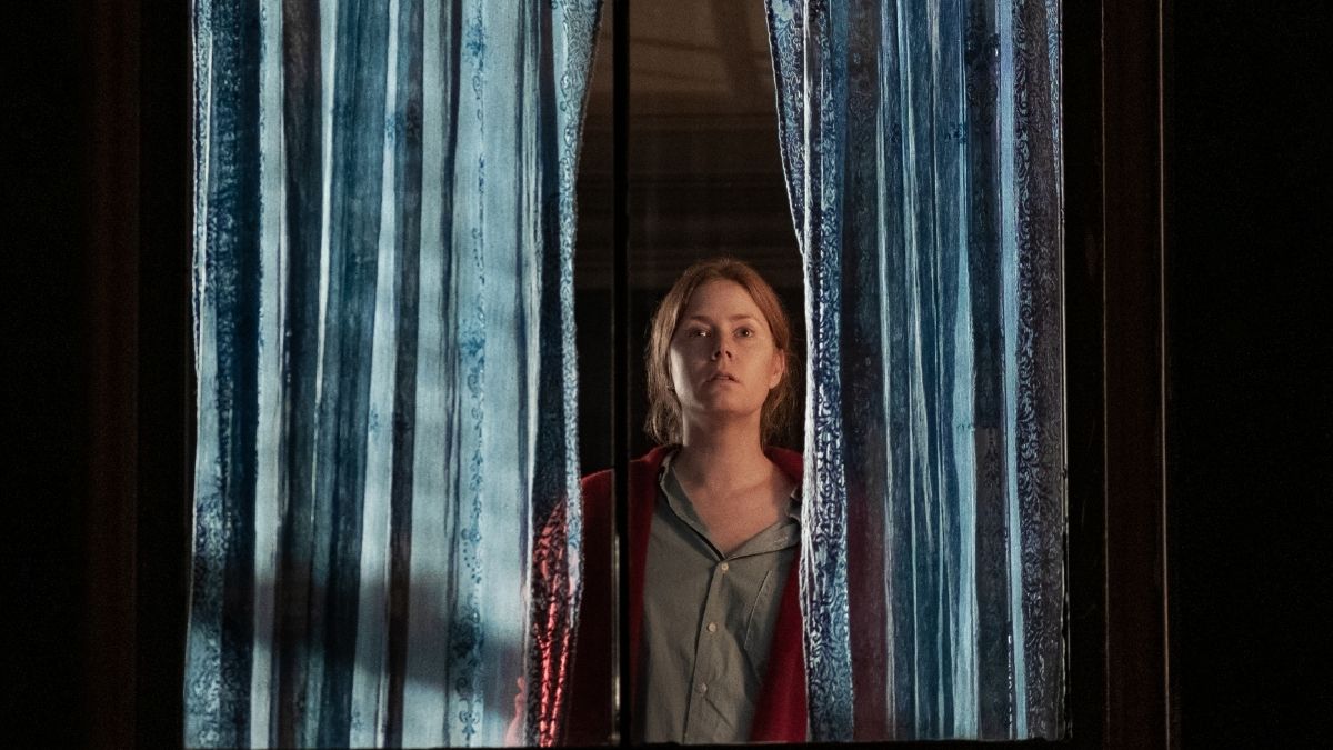 Cena do filme A Mulher na Janela. Nela, vemos Amy Adams, branca, ruiva, de camiseta cinza e jaqueta vermelha, olhando pela janela, com duas cortinas dividindo a imagem e ela no centro, com uma expressão aflita.