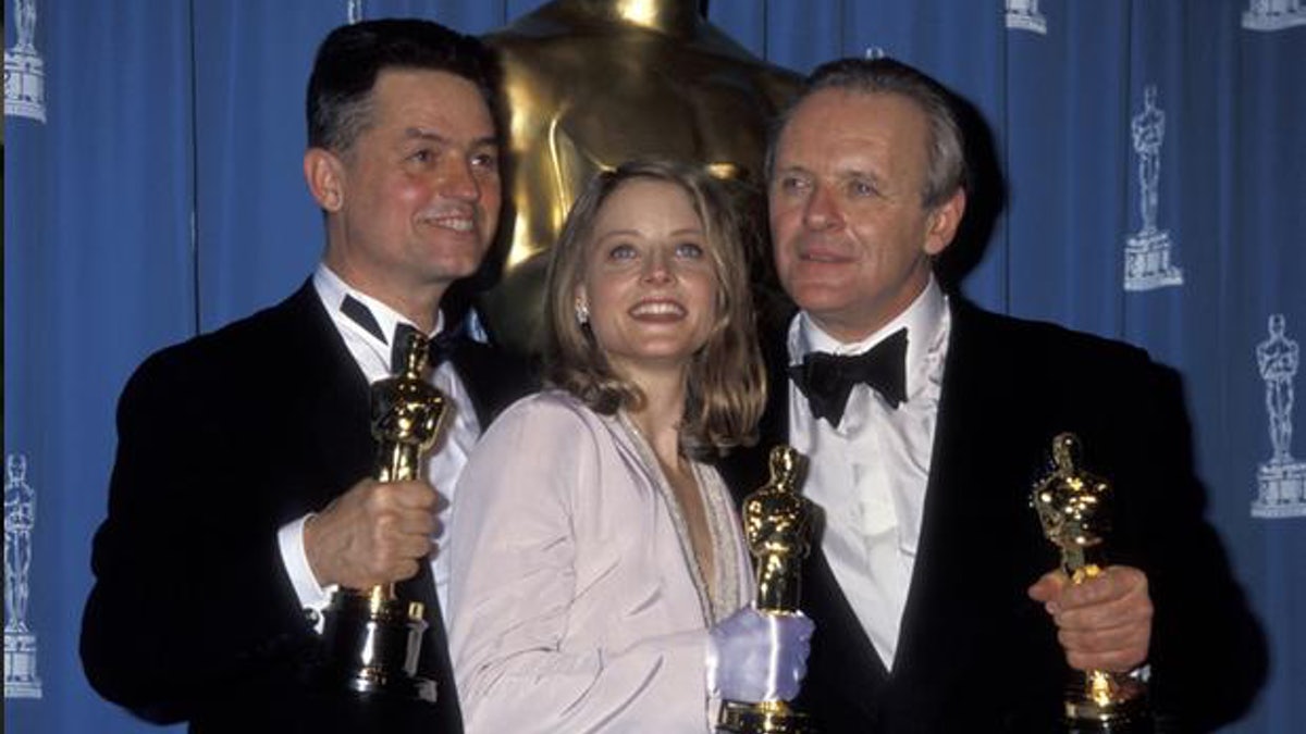 Foto de três vencedores do Oscar. Jonathan Demme, Jodie Foster e Anthony Hopkins estão de pé, cada um segurando seu Oscar, uma estatueta dourada e comprida no formato de um homem. Os dois homens usam ternos pretos com blusas brancas, enquanto Jodie usa um terno bege claro. Os três sorriem.