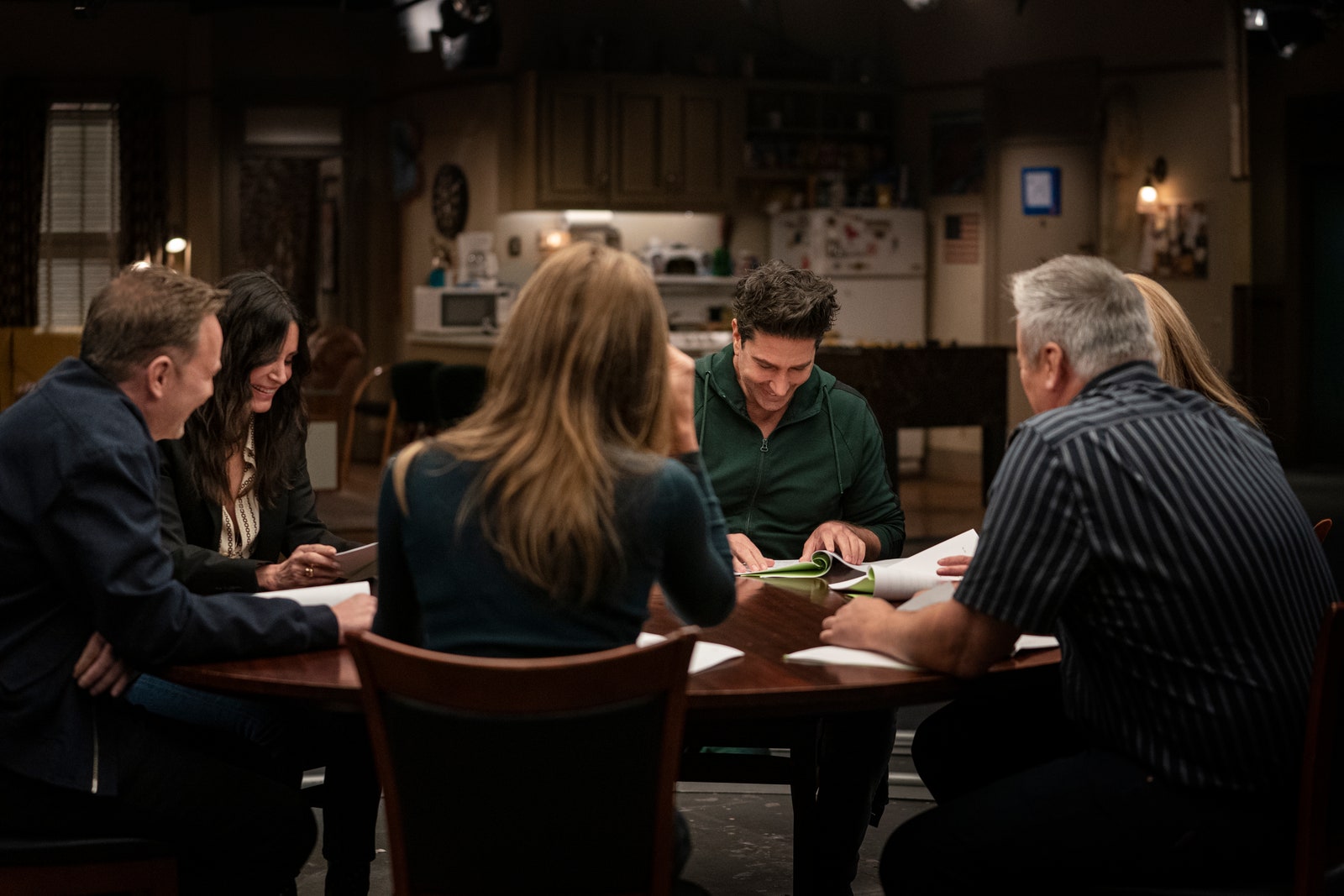 Cena do episódio especial Friends: The Reunion. Nela, vemos os 6 atores sentados ao redor de uma mesa de madeira, com roteiros de papel sulfite na mão, rindo e se olhando. 