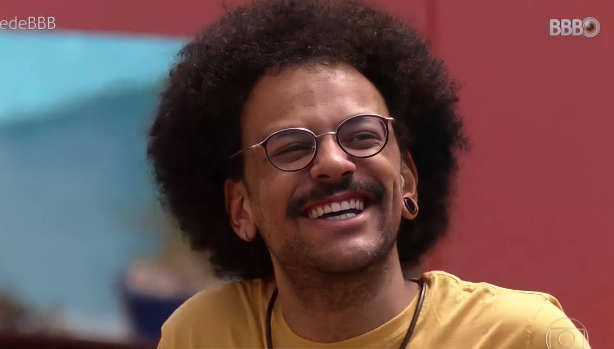 Cena do Big Brother Brasil 21. João Luiz, vestindo uma camiseta laranja e óculos, sorri. 